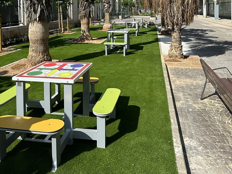 Un área ajardinada con mesas y bancos con juegos clásicos como parchis, ajedrez